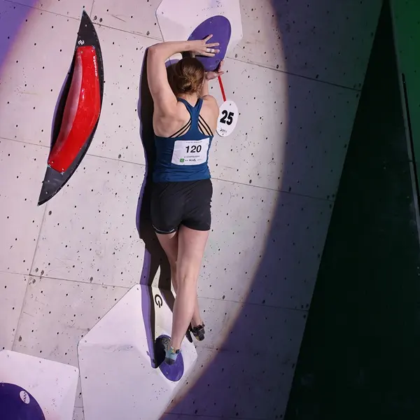 Een vrouwelijke atlete in een tanktop en korte broek beklimt een indoor-bouldermuur; ze strekt haar rechterarm omhoog en drukt haar linkerarm tegen een grote paarse greep met het nummer 25 erop. Ze draagt klimschoenen en het startnummer 120.